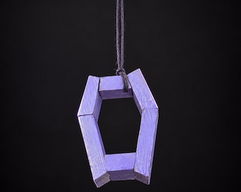 Puede incluir: Un adorno en forma de ataúd de color violeta claro, construido con seis segmentos de madera individuales. Las piezas están unidas para formar un marco geométrico hueco. Un cordel gris oscuro pasa por el segmento superior para colgarlo.