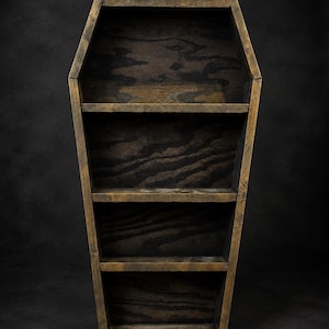 May include: A coffin-shaped wooden shelf unit featuring a dark, stained finish with visible wood grain patterns. The structure includes three horizontal shelves within the tapered frame, providing four distinct storage sections for display purposes.