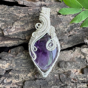 Può includere: Una gemma di ametista viola a forma di goccia, incastonata in una montatura in filo metallico color argento. Il filo è avvolto e intrecciato in modo complesso attorno alla pietra, formando una cornice decorativa che termina con un occhiello.