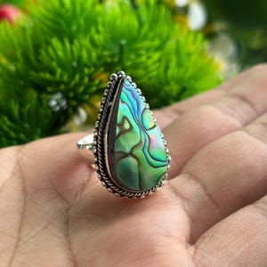 Puede incluir: Un anillo en forma de lágrima con una incrustación de concha de abulón iridiscente en tonos verdes, azules y morados. La piedra está engastada en un marco de metal plateado con un borde decorativo de cuerda trenzada y una banda lisa.