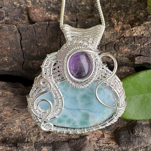 May include: A wire-wrapped pendant featuring a large, smooth, light blue stone at the base and a smaller, oval-shaped purple stone set above it. The silver-toned wire is intricately coiled around both stones, forming a decorative frame.