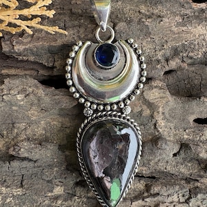 Puede incluir: Un colgante de plata con una piedra oscura en forma de lágrima con inclusiones verdes en la base, rematado por un elemento de plata en forma de media luna y una pequeña piedra azul oscuro facetada. El colgante incluye una reasa de plata.