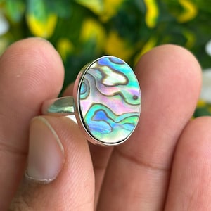 May include: A silver ring featuring an oval-shaped abalone shell inlay. The shell displays a vibrant, iridescent surface with swirling patterns of blue, green, pink, and purple hues. The metal band is smooth, polished, and silver in color.
