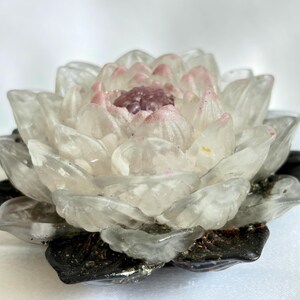 May include: A detailed, clear glass lotus flower sculpture. The petals are white and translucent with pink tips, surrounding a darker pink center. The base is dark gray and black.