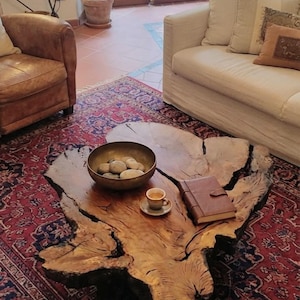 Può includere: Un tavolino da salotto in legno con bordo naturale, venature a vista e bordi scuri irregolari. Sulla superficie si trovano una ciotola metallica con pietre lisce, una tazzina in ceramica con piattino e un diario in pelle marrone chiuso.
