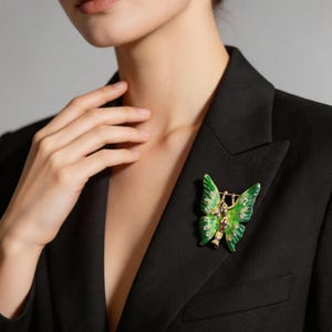 May include: A green and gold butterfly brooch with a small figure in the center, set with small crystals. The brooch is pinned to a black blazer.