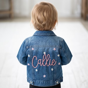 May include: A blue denim jacket featuring the name Callie embroidered in pink script across the back. The jacket is decorated with small, scattered embroidered flowers in shades of pink and white, and includes a classic collar and buttoned cuffs.