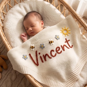 Op de afbeelding: Een crèmekleurige gebreide babydeken met de naam Vincent geborduurd in roestkleurig garen. De deken is versierd met kleine, verhoogde applicaties van twee bijtjes, drie lichtblauwe bloemen en een gele zon, allemaal in een gebreide textuur.