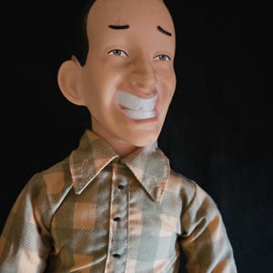 May include: A doll with a molded head featuring a prominent pointed hairstyle, arched eyebrows, and a wide, toothy grin. The figure wears a long-sleeved button-down shirt with a tan and olive green plaid pattern and a dark-colored lower garment.