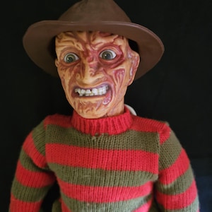 May include: A figure of a character with a scarred, textured face, wearing a brown fedora hat and a long-sleeved sweater featuring horizontal red and olive green stripes. The figure has wide, light-colored eyes and bared teeth, with a knit texture on the sweater fabric and a smooth, molded appearance on the facial features.