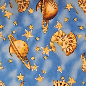 May include: A blue fabric featuring a repeating pattern of golden yellow celestial bodies including planets with rings, suns with faces, and various sizes of five-pointed stars. The background has a mottled, cloud-like texture in shades of blue.