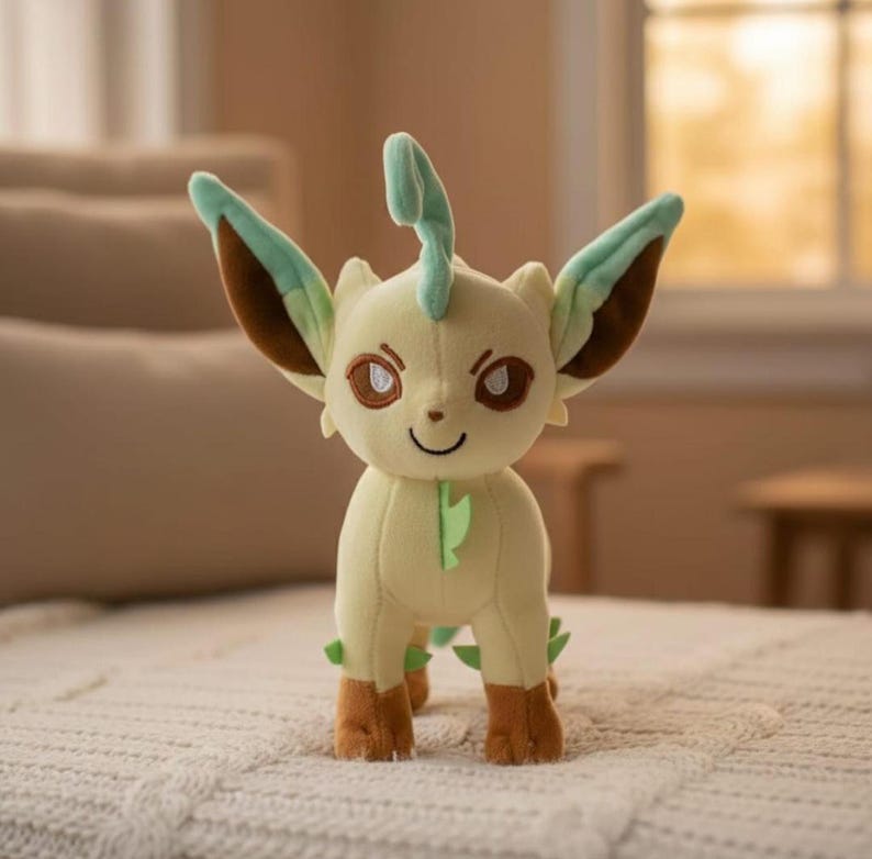 May include: A plush toy of a Leafeon Pok&eacute;mon character. The plush has a light yellow body, brown and green accents on the ears and feet, and a smiling face. The toy is standing on a white surface.