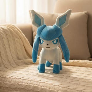 May include: A plush Glaceon toy in shades of blue and white. The toy features large, pointed ears with diamond-shaped accents, long, flowing appendages, and a friendly expression. It is posed on a cream-colored blanket.