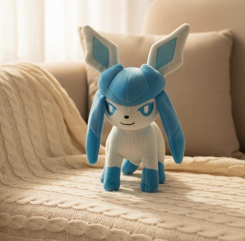 May include: A plush Glaceon toy in shades of blue and white. The toy features large, pointed ears with diamond-shaped accents, long, flowing appendages, and a friendly expression. It is posed on a cream-colored blanket.