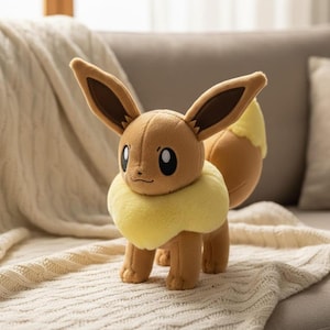 May include: A plush Eevee toy, featuring brown fur, large ears with dark brown inner ears, and a fluffy yellow collar. The toy has a small, smiling face with large, oval eyes and a fluffy yellow tail. It is standing on a cream-colored blanket.