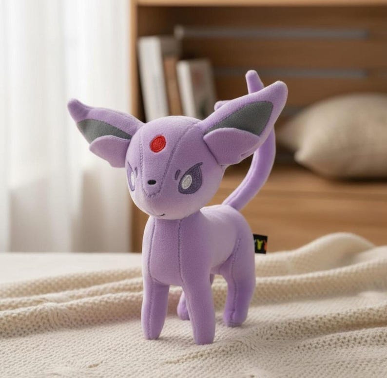 May include: A plush toy of a purple creature with large ears, a long tail, and a red spot on its forehead. The toy has gray accents on its ears and is standing on a cream-colored blanket. The toy is a popular character.