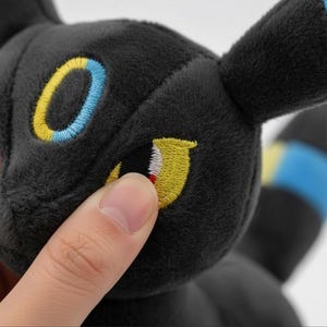 May include: A black plush toy with embroidered yellow, blue, and red details. The toy has large, expressive eyes and a soft, textured surface. The design is reminiscent of a popular animated character.