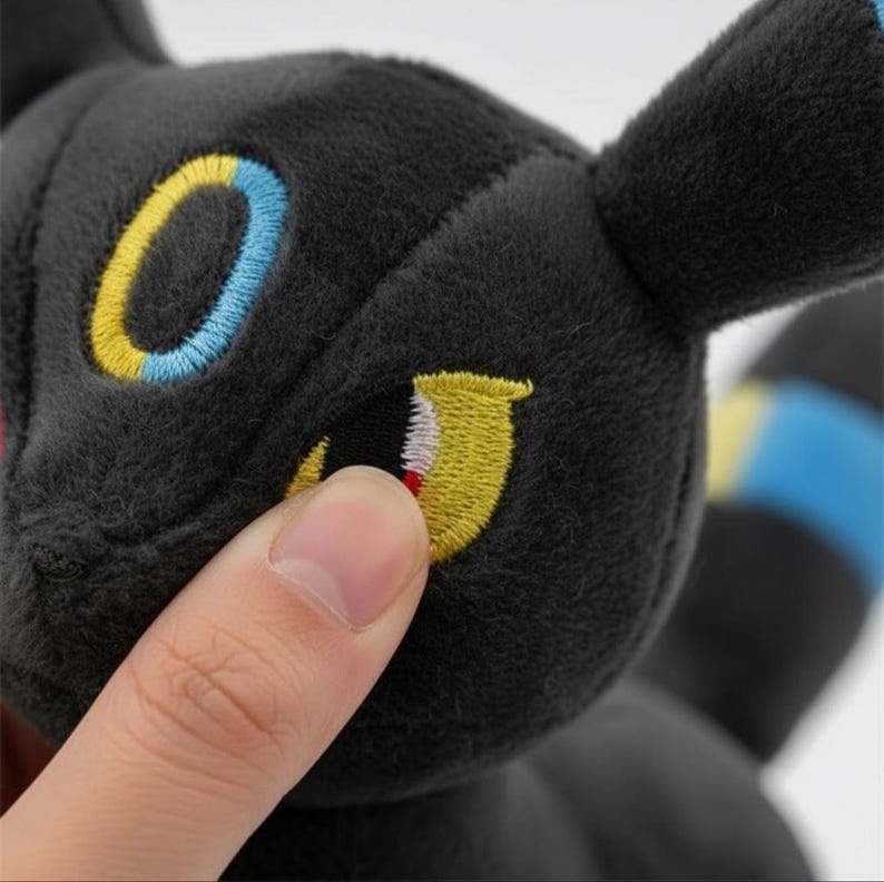 May include: A black plush toy with embroidered yellow, blue, and red details. The toy has large, expressive eyes and a soft, textured surface. The design is reminiscent of a popular animated character.