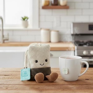 Puede incluir: Un caprichoso personaje de bolsa de té de peluche con una cara sonriente, pies marrones y una etiqueta azul claro, junto a una taza blanca de té con una bolsita de té. La escena está ambientada sobre una superficie de madera en una cocina.