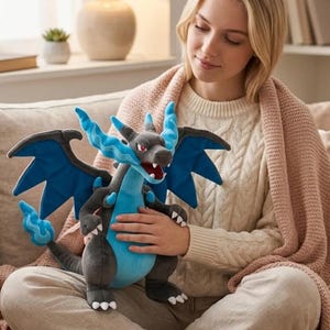 May include: A plush toy dragon in shades of gray, blue, and red. The dragon has large wings, a long tail, and a detailed face. The toy is being held by a person wearing a cream-colored sweater.
