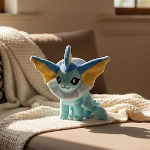 May include: A plush toy of a blue and yellow creature with large ears and a fin. The toy has a white collar and a small tail. It is sitting on a cream-colored blanket.