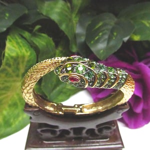 May include: A gold-toned hinged bracelet shaped like a snake. The body features a textured scale pattern, while the head is encrusted with numerous small green crystals and two prominent red crystals serving as eyes, creating a detailed finish.