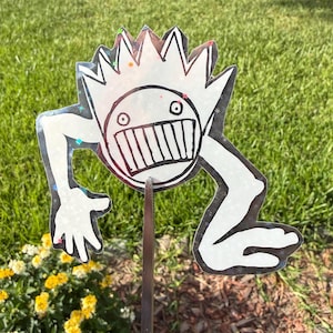 May include: A whimsical, white and black cartoon character yard stake with a spiky crown, large grin, and outstretched arms and legs. The character is mounted on a metal stake, set against a backdrop of green grass and yellow flowers.