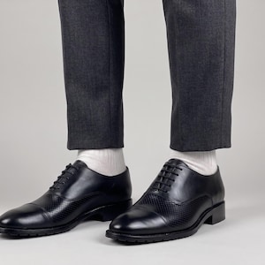 May include: A pair of black leather oxford shoes featuring a cap toe design with perforated detailing on the vamp and sides. The shoes have black laces, a low heel, and a textured rubber outsole, worn with white socks and charcoal gray trousers.