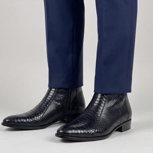 Könnte beinhalten: Ein Paar schwarze Stiefeletten mit strukturiertem Schlangenledermuster, spitzer Zehenpartie und seitlichem Reißverschluss, getragen zu einer dunkelblauen Hose. Die Stiefel haben einen niedrigen Blockabsatz und eine glatte, polierte Oberfläche.