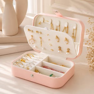 May include: A pink jewelry box with multiple compartments, open to display various gold earrings, rings, and necklaces. The interior is lined with white fabric. The box is closed with a small clasp.