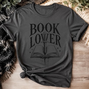 Könnte beinhalten: Dunkelgraues T-Shirt mit einem grafischen Design. Das Design zeigt die Worte "BOOK LOVER" über einem offenen Buch, das von einem Schwert durchbohrt wird, mit Sternen und einem Tropfeffekt. Das T-Shirt ist unten geknotet.