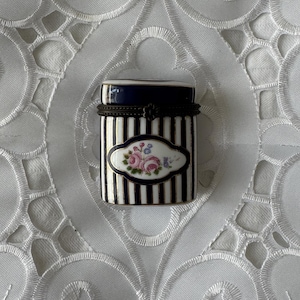 Puede incluir: Una pequeña caja de porcelana cilíndrica con tapa azul oscuro y cuerpo decorado con rayas verticales negras y blancas. Un medallón ovalado central en la parte frontal muestra un motivo floral pintado a mano con rosas rosadas y hojas verdes.
