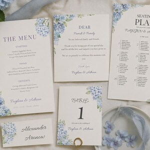 May include: A collection of wedding stationery featuring a menu, seating plan, and table number cards. The cards are decorated with blue hydrangea floral arrangements. The menu includes starters, mains, and desserts. The seating plan lists names. The stationery is for Faylinn & Adrian.