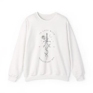 May include: White sweatshirt with a graphic design featuring a sword, rose, and crescent moon. The text on the sweatshirt reads "IN LOVE WITH MORALLY GREY MEN."