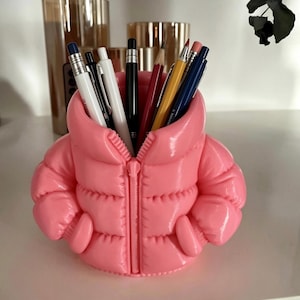 May include: A bright pink pencil holder shaped like a miniature puffer jacket with a central zip detail and two side pockets. The jacket is open at the top, serving as a container for various pens, pencils, and markers of different colours.