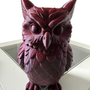 May include: A dark maroon owl figurine featuring detailed moulded feather textures across its body, pointed ear tufts, and large circular eyes. The sculpture is rendered in a solid, deep red-purple tone with a matte finish and visible layered plumage.