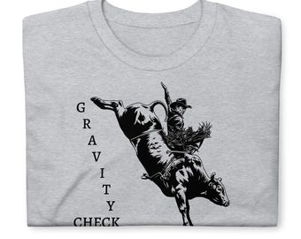 May include: A light grey t-shirt featuring a black graphic illustration of a cowboy riding a bucking bull. The text "GRAVITY CHECK" is printed vertically in black capital letters to the left of the bull rider illustration on the front.