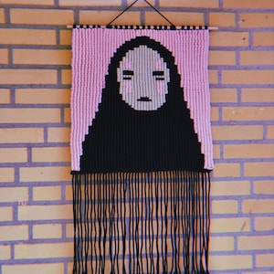 May include: A macrame wall hanging featuring a black and pink design. The artwork depicts a character with a black cloak and a pale face with pink cheeks. The piece is rectangular with a fringe at the bottom and is suspended from a wooden dowel.