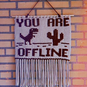 May include: Macrame wall hanging with the words "YOU ARE OFFLINE" in dark brown. A dinosaur and cactus are featured between the words. The wall hanging is beige with a fringe at the bottom and is suspended from a wooden dowel.