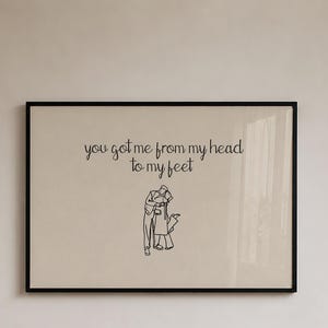 May include: A rectangular wall art print in a black frame featuring a beige background with black line art of two people embracing and the text "you got me from my head to my feet" written in a cursive black font centered above the illustration.