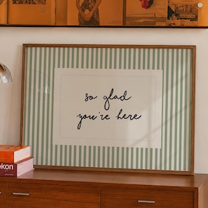 Puede incluir: Un revistero de pared de acrílico naranja con cuatro revistas, una lámpara de mesa de cromo en forma de hongo, dos libros apilados y una lámina enmarcada con rayas verticales verdes y el texto "so glad you're here" sobre un aparador de madera.
