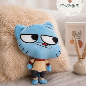 May include: A plush blue cartoon cat doll with large white-rimmed eyes and a neutral expression. The doll wears a tan shirt with a red collar and cuffs, gray shorts, and blue shoes. The doll is on a fluffy beige pillow.