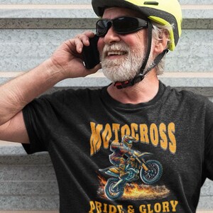 May include: A charcoal gray t-shirt featuring a graphic print of a motocross rider on a motorcycle with flames. The text reads MOTOCROSS PRIDE & GLORY EST. 1952 in yellow and white lettering. The shirt is worn by a person with a yellow helmet and sunglasses.