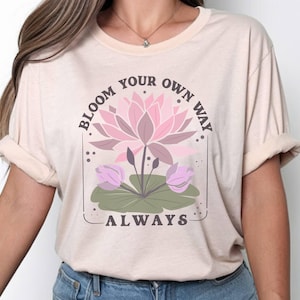 May include: A light beige short-sleeve t-shirt featuring a graphic of a pink lotus flower with green lily pads. The text "BLOOM YOUR OWN WAY" is printed in a dark brown arched font above the flower, and the word "ALWAYS" is printed below it.