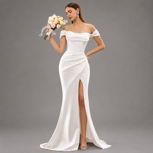 May include: A white off-the-shoulder wedding dress with a high slit. The gown has a fitted bodice and a flowing skirt. The bride is holding a bouquet of pink and white flowers.