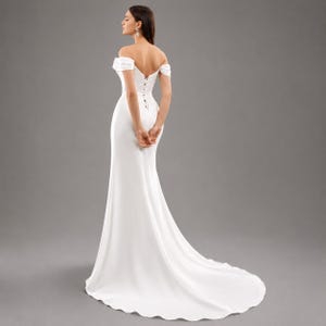 May include: A white off-the-shoulder wedding dress with a fitted bodice and a flowing skirt. The dress has a subtle train and a delicate button detail down the back. The model is standing with their back to the camera.