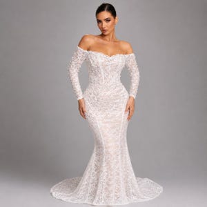 May include: A white, off-the-shoulder wedding dress with long sleeves. The gown has detailed lace and a mermaid shape, with a fitted bodice and a flared skirt that flows into a train. The sleeves are decorated with small, textured embellishments.