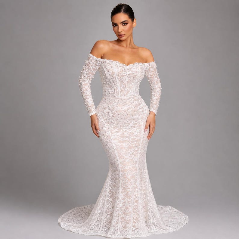 May include: A white, off-the-shoulder wedding dress with long sleeves. The gown has detailed lace and a mermaid shape, with a fitted bodice and a flared skirt that flows into a train. The sleeves are decorated with small, textured embellishments.
