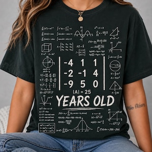 May include: A dark charcoal gray short-sleeve t-shirt featuring a white graphic print of various mathematical formulas, geometric shapes, and a central matrix calculation with the text 25 YEARS OLD printed in a bold white font below it.