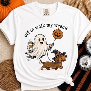 Puede incluir: Una camiseta blanca de manga corta con un gráfico de un fantasma que sostiene una taza de café y un globo de calabaza, paseando a un perro salchicha con sombrero de bruja. El texto sobre el gráfico dice, off to walk my weenie.